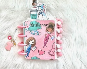May include: A pink and white planner with a pom-pom trim. The cover features a cartoon nurse with a stethoscope and a heart. The planner is personalized with the name "Kristianne".