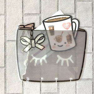 May include: A clear plastic pocket with a gray background and a white bow. The pocket has a white eye design and the text "Sandys Planner Shop" and "Coffee Lover" are visible.