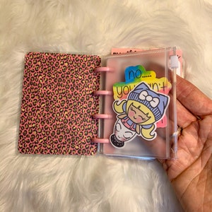 Peut inclure: Un carnet à couverture léopard rose et or avec une poche transparente en plastique sur le côté droit. La poche contient un autocollant représentant une fille de dessin animé avec le texte "no... you won't".