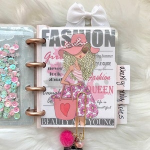 May include: A pink and white planner with a fashion-themed design featuring a girl with a pink hat and a pink purse. The planner has a gold ring binder and a gold clip with a pink charm. The planner has the words "Fashion Queen" and "Beauty and Young" printed on it.