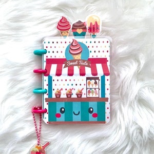May include: A pink and blue ice cream shop themed notebook with a colorful polka dot design. The notebook has a clear plastic cover with a cute ice cream cone design. The notebook has a metal ring binder and a pink charm with a cute ice cream cone design.