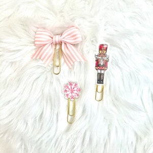 May include: Three decorative paper clips on a white fluffy surface. One clip features a pink and white striped bow, another a pink snowflake, and the third a nutcracker design. All clips have gold-colored metal clasps.