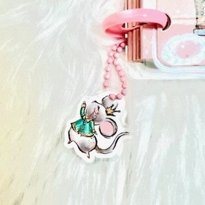 May include: A pink and white charm with a cartoon mouse wearing a green and gold outfit and a crown. The charm is attached to a pink chain.