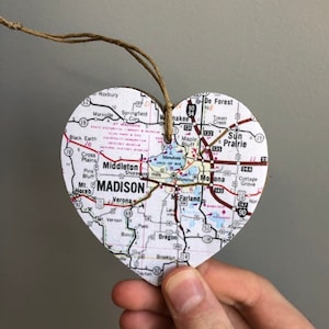 Puede incluir: Un adorno en forma de corazón hecho de madera con un mapa de Madison, Wisconsin impreso en él. El mapa muestra las calles, autopistas y pueblos circundantes de la ciudad. El adorno está colgado de una cuerda de yute marrón.