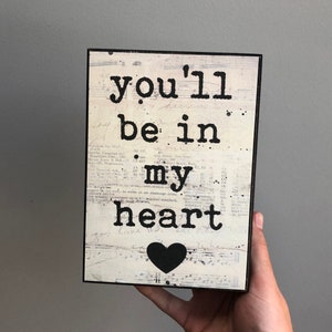 Może przedstawiać: Drewniany znak z białym tłem i czarnym tekstem, który brzmi "you'll be in my heart". Pod tekstem znajduje się czarne serce.