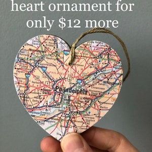 May include: A heart-shaped ornament made of wood with a map of Philadelphia printed on it. The ornament is hanging from a string and is being held by a hand. The text "Add a Philadelphia heart ornament for only $12 more" is visible at the top of the image.