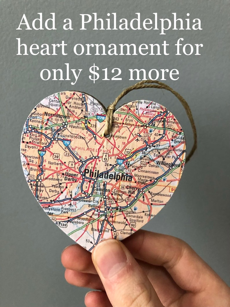 May include: A heart-shaped ornament made of wood with a map of Philadelphia printed on it. The ornament is hanging from a string and is being held by a hand. The text "Add a Philadelphia heart ornament for only $12 more" is visible at the top of the image.
