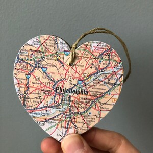 May include: A heart-shaped wooden ornament with a map of Philadelphia, Pennsylvania printed on it. The ornament is decorated with red, blue, and green lines representing roads and highways. The ornament is hanging from a brown twine string.