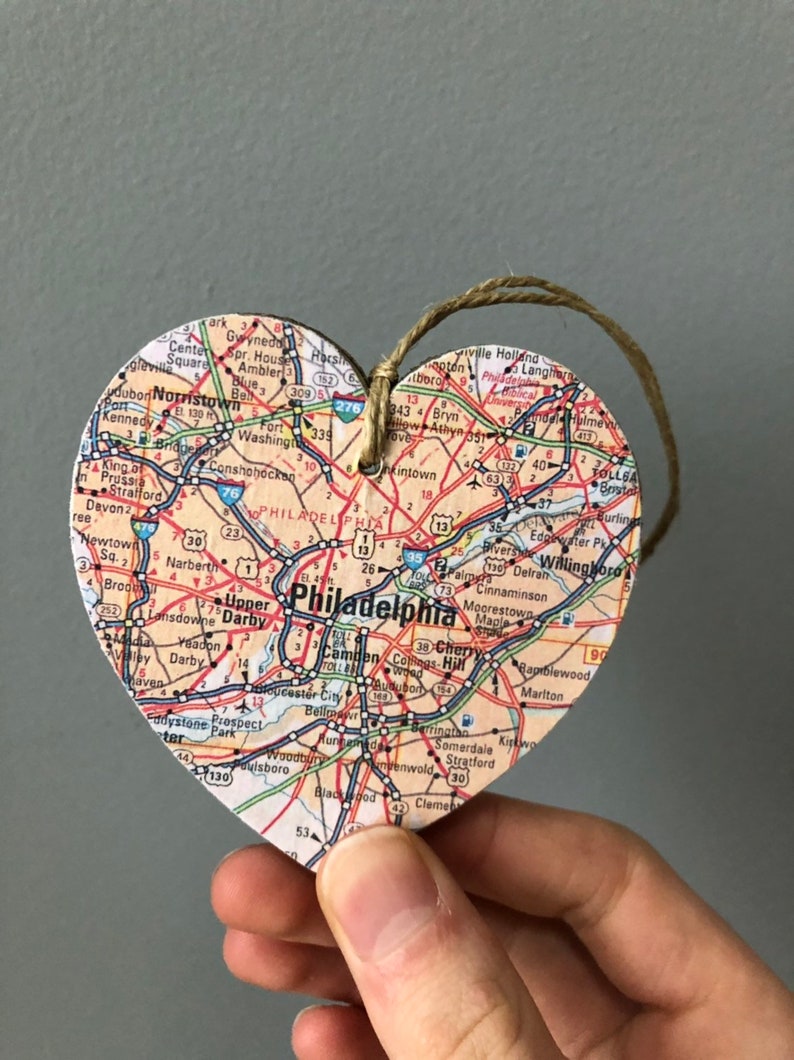 May include: A heart-shaped wooden ornament with a map of Philadelphia, Pennsylvania printed on it. The ornament is decorated with red, blue, and green lines representing roads and highways. The ornament is hanging from a brown twine string.