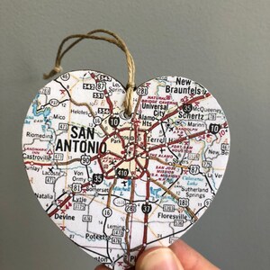 May include: Heart-shaped wooden ornament featuring a map of San Antonio, Texas. The map includes roads, landmarks, and city names. A twine loop is attached for hanging. The ornament is held by a hand.