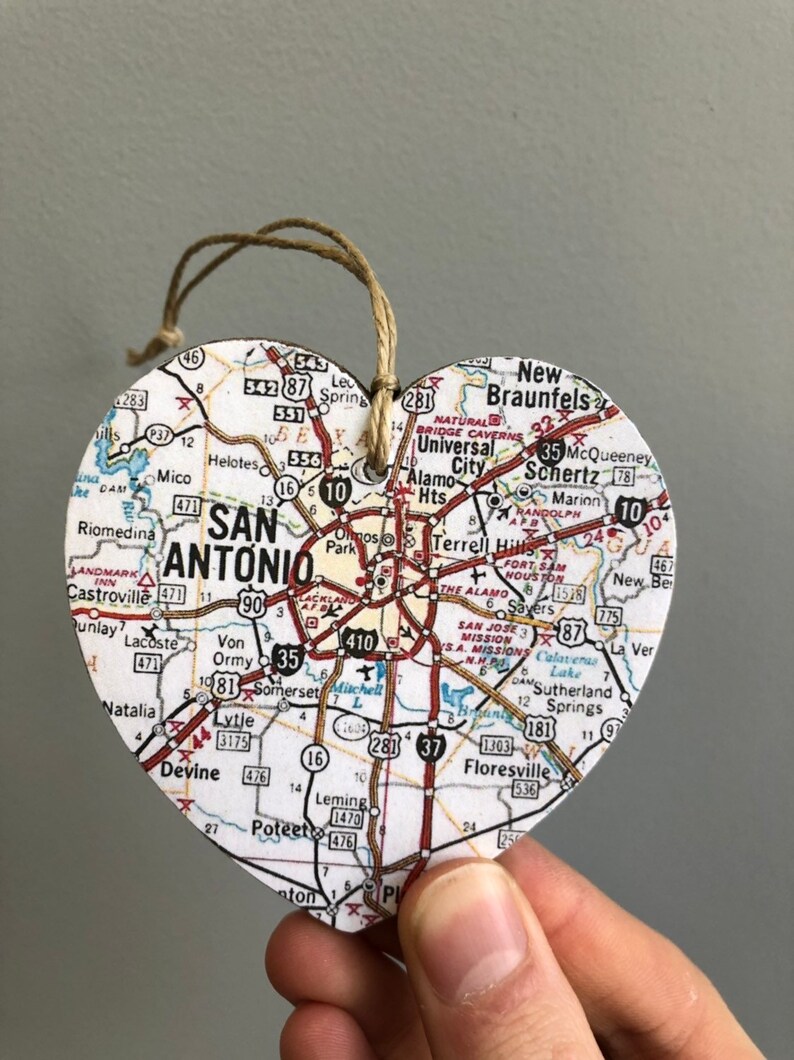 May include: Heart-shaped wooden ornament featuring a map of San Antonio, Texas. The map includes roads, landmarks, and city names. A twine loop is attached for hanging. The ornament is held by a hand.