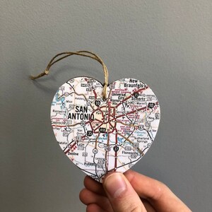 May include: A heart-shaped ornament featuring a map of San Antonio, Texas. The map includes city streets, highways, and surrounding towns. A twine loop is attached for hanging. The ornament is held by a hand.