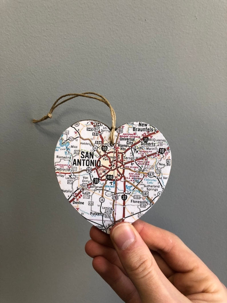 May include: A heart-shaped ornament featuring a map of San Antonio, Texas. The map includes city streets, highways, and surrounding towns. A twine loop is attached for hanging. The ornament is held by a hand.