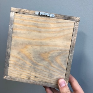 May include: The back of a wooden picture frame with a metal hanging hook. The frame is made of natural wood with a light brown finish.