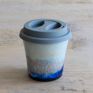 May include: A ceramic travel mug with a light blue and brown glaze, topped with a gray silicone lid. The mug has a cylindrical shape and a black base. The lid has two openings and the text "CAUTION HOT" is visible on the lid.