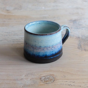 Puede incluir: Una taza de cerámica hecha a mano con forma redondeada y un asa pequeña. La taza presenta un degradado de colores, que va del azul claro interior a una base azul oscuro. El borde tiene un acento marrón.