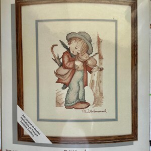 May include: Counted cross-stitch kit titled "Needle Treasures" featuring a framed cross-stitch of "The Little Fiddler." The artwork depicts a child playing a violin, with an umbrella and a building in the background. The frame size is 12 x 14 inches.