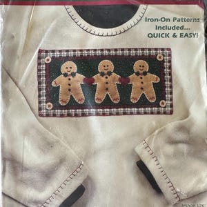 May include: A craft kit with felt and fabric appliqués. The design features three gingerbread figures with red hearts and bow ties. The kit includes iron-on patterns and measures approximately 28 cm x 15 cm.
