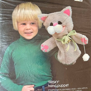 May include: A Bucilla kit featuring a gray plush cat hand puppet with pink accents, a green bow, and a white pom-pom. The kit includes all materials and instructions. The packaging shows a child wearing a green turtleneck.
