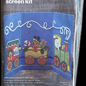 Puede incluir: Kit de fieltro empaquetado para una pantalla con temática navideña. El kit presenta un fondo azul con un diseño de tren de fieltro. Los vagones están decorados con Papá Noel, renos y regalos. Tamaño final: 56,5 x 35,6 cm.