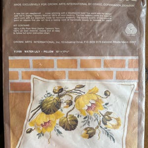 May include: A Count-A-Point needlecraft kit by Crown Arts. The kit includes a pillow with a Water Lily design, measuring 15" x 17.5". The pillow features a floral cross-stitch pattern in yellow, green, and brown on a white background.