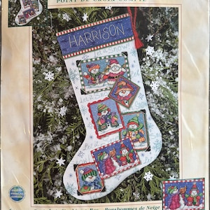 May include: A Dimensions counted cross stitch kit featuring a Christmas stocking with a snowman theme. The stocking is white with blue accents and the name "HARRISON". The design includes multiple framed images of snowmen, with the text "Let it Snow!". The stocking is approximately 41 cm long.