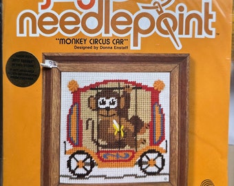 May include: A Jiffy Needlepoint kit featuring a "Monkey Circus Car" design. The kit includes a needlepoint canvas with a framed image of a monkey in a circus car, with the text "MONKEY CIRCUS CAR" and "Designed by Donna Enstaff". The package is orange.
