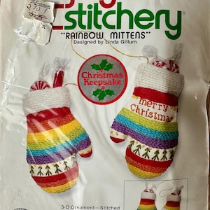 Puede incluir: Un kit de manualidades Jiffy Stitchery para adornos Rainbow Mittens. El paquete muestra dos manoplas a rayas coloridas con puños blancos, patrones de árboles verdes y el texto Merry Christmas. Incluye una etiqueta Christmas Keepsake.