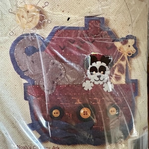 May include: A Feltworks applique kit featuring a Noah's Ark design. The kit includes felt, buttons, and embroidery thread. The ark is red and blue, with felt animal figures. The package size is 8" x 8".