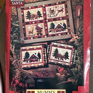 May include: A red and brown Christmas-themed quilt pattern with the text "WOODLAND SANTA" at the top. The quilt design features squares with images of Santa, bears, trees, and cabins. The bottom of the image has the text "MUMM'S THE WORD".