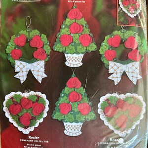 Puede incluir: Un kit de adornos de fieltro Bucilla con un diseño de rosal. El kit incluye seis piezas, con fieltros estampados, hilo de algodón, lentejuelas, cuentas e hilo metálico. Los adornos incluyen coronas de rosas, formas de corazón y formas de árbol. Diseñado por Maria Stanzioni.