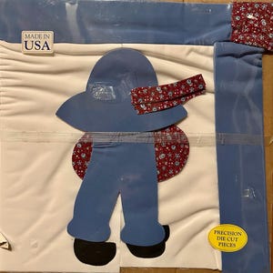 May include: A craft kit featuring a blue figure with a hat, pants, and a floral patterned accent. The kit includes the text "MADE IN USA" and "PRECISION DIE CUT PIECES". The figure is on a white background.