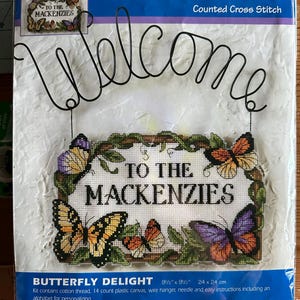 May include: A Wire Welcome counted cross stitch kit featuring a black wire hanger with the word Welcome and a plastic canvas design of butterflies and vines surrounding the text To The Mackenzies. The package measures 9.5 inches by 9.5 inches.
