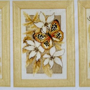 May include: Three framed needlepoint artworks featuring butterflies and flowers. The butterflies have orange, black, and blue wings. The flowers are white. The frames are a light beige color. The artwork is displayed on a beige background.