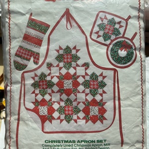 May include: A Christmas Quickit apron set in a plastic package. The set includes a pre-printed apron, mitt, and two potholders with a red, white, and green Christmas design. The apron is approximately 23 inches long, and the mitt is 12 inches.
