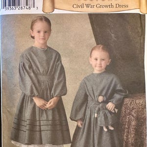 May include: Two young girls wearing Civil War Growth Dresses. The dresses are gray with long sleeves, a fitted bodice, and a skirt that falls to the mid-calf. The dresses have white lace trim at the hem. The image includes the text "Simplicity Historic Costumes Civil War Growth Dress".