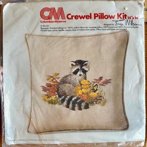 May include: A Columbia-Minerva crewel pillow kit in original plastic packaging. The kit features a 14" x 14" stamped design of a raccoon sitting among autumn leaves. The packaging includes the brand name, Erica Wilson design credit, and contents.