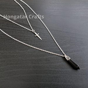 Könnte beinhalten: Mehrschichtige Halskette in Silberoptik mit einem kleinen Dolch-Anhänger und einem schwarzen, spitz zulaufenden Kristall-Anhänger. Die Halskette wird auf einer dunklen Oberfläche gezeigt. Der Text "Hongata Crafts" ist sichtbar.