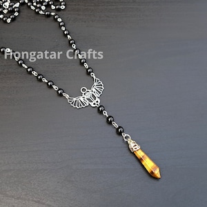 Puede incluir: Un collar de cuentas de ónix negro con un colgante de plata con un diseño de escarabajo alado. El colgante está unido a un largo cristal de ojo de tigre puntiagudo.