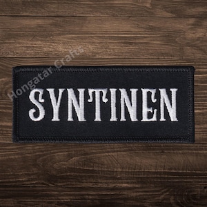 May include: A black rectangular patch with the word "SYNTINEN" embroidered in white capital letters. The patch is likely made of fabric and has a stitched border. The background is a dark wood surface.
