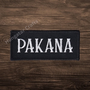 Pode incluir: Um patch retangular preto com a palavra "PAKANA" bordada em branco. O patch tem uma borda preta e está sobre um fundo de grão de madeira escura. O texto está em negrito.