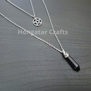 Puede incluir: Un collar de cadena de plata con dos colgantes. El colgante superior es un pentagrama de plata. El colgante inferior es una punta de cristal negro.