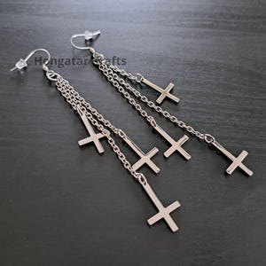 Può includere: Orecchini pendenti color argento con ciondoli a croce rovesciata. Ogni orecchino ha più catene con croci, creando un effetto a strati. Gli orecchini sono esposti su una superficie scura.
