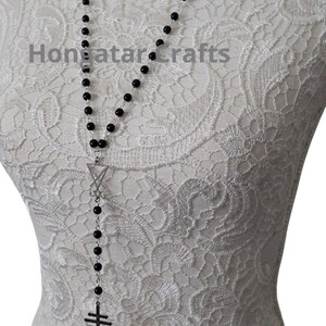 May include: A long black beaded necklace with a silver pentagram pendant and a black symbol pendant. The necklace is displayed on a white mannequin with a lace pattern. The text "Hongatar Crafts" is visible.