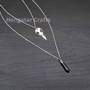 May include: A silver layered necklace with two pendants. The top pendant is a silver bird skull, and the bottom pendant is a black gemstone.