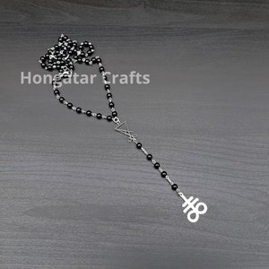 May include: A black beaded necklace with a silver-tone pendant featuring a stylized upside-down triangle and a symbol. The necklace is displayed on a dark gray wooden surface. The text "Hongatar Crafts" is visible.