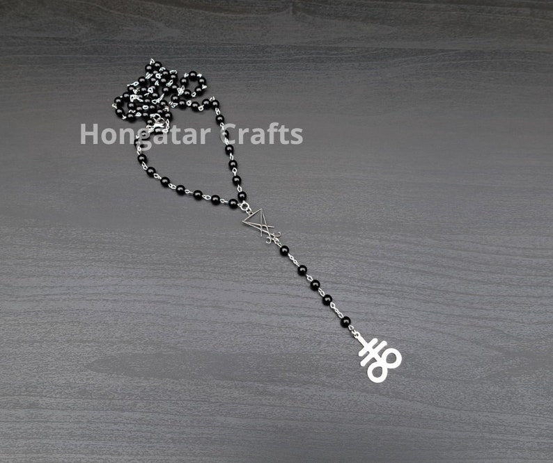 May include: A black beaded necklace with a silver-tone pendant featuring a stylized upside-down triangle and a symbol. The necklace is displayed on a dark gray wooden surface. The text "Hongatar Crafts" is visible.