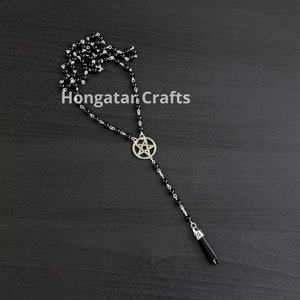 Puede incluir: Un collar de cuentas negras con un colgante de pentagrama plateado y una punta de cristal negro en el extremo. El collar está sobre una superficie de madera oscura. "Hongatar Crafts" está escrito en blanco sobre la superficie.