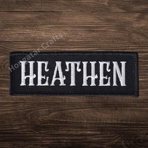 May include: A black rectangular patch with the word "HEATHEN" embroidered in white capital letters. The patch is set against a dark wooden background.