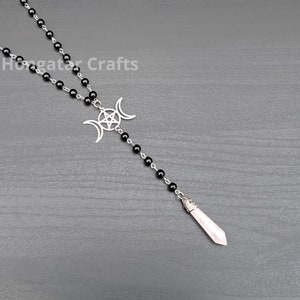 Puede incluir: Un collar de cuentas negras con un colgante de triple luna plateado y una punta de cuarzo rosa. El colgante presenta un pentagrama dentro de las lunas.